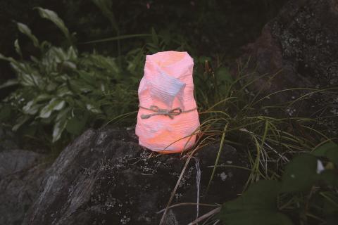 A handmade, orange, lantern-like object with a bow is sitting on a rock in a natural outdoor setting.