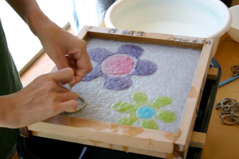 A person's hands are working on a wooden frame, creating a flower design with colored paper pulp submerged in water.