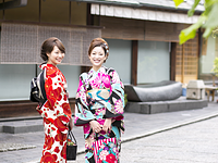 Two smiling women in vibrant, floral kimonos stand on a street in front of a traditional Japanese building.