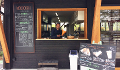 A smiling man stands in a food stall, advertising NOQOO DERI with a menu of food, drinks, and desserts like hot dogs, coffee, cider, and soft cream, showcasing a "Today's menu" sign.