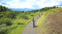 A group of cyclists ride along a paved path in a lush, green landscape with mountains in the distance.