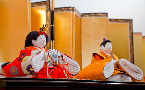A photograph captures two traditional Japanese Hina dolls adorned in vibrant orange and red garments, seated against a backdrop of golden folding screens.