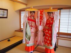 Two women in traditional Japanese wedding attire, including tall straw hats and veils, stand in a room with tatami mats and shoji screens, posing with smiles.