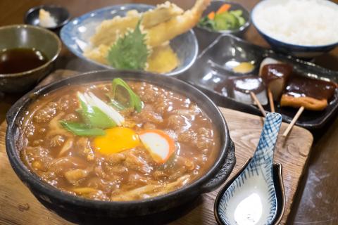 A steaming bowl of Japanese noodle soup with green onions, egg yolk, and fish cake is the centerpiece of this food spread, complemented by tempura, dango skewers, rice, and dipping sauce.