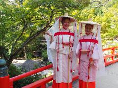 Two women in traditional Japanese attire, including straw hats with veils and red accents, stand on a red bridge surrounded by lush greenery.