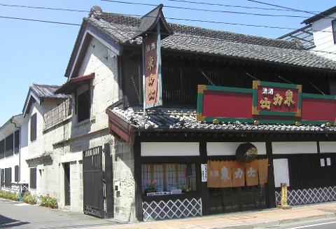 A two-story traditional Japanese building with gray tiled roofs, featuring signs with Japanese writing, fabric curtains, and intricate lattice details.