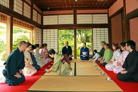 A group of people are seated around a tatami mat during a traditional Japanese tea ceremony.
