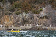 Two kayakers paddle along a rocky coastline with a white fence and steps leading up a cliff face covered in vegetation.