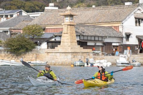 Two people are kayaking in a calm bay with traditional Japanese buildings and a stone lantern in the background.