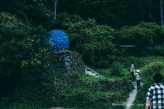 "Reborn-Art" Tour: Experience Contemporary Art on the Oshika Peninsula A blue, spherical sculpture is displayed amidst lush greenery and stone walls, with three people walking on a path nearby.