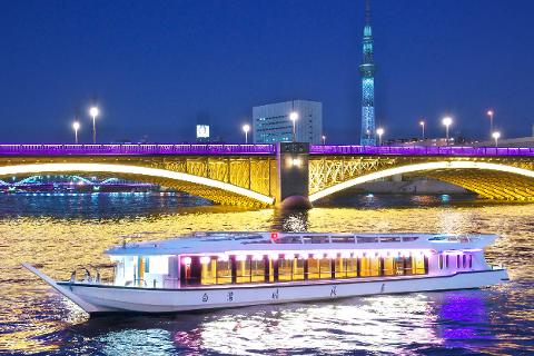 A boat is illuminated in front of a bridge in Tokyo at night, with the Tokyo Skytree visible in the background.
