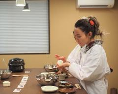 A woman in traditional Japanese attire prepares food in a room with a window, various bowls, and cooking utensils on a table.