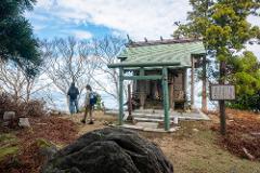 Two tourists admire a shrine at the top of Kinkasan Island, surrounded by trees and overlooking the sea.