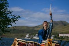 A Japanese man in traditional attire plays a taiko drum beside a lake, his stick raised in the air against a backdrop of rolling hills.
