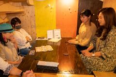 A group of four people, two women on each side, sits around a wooden table in what appears to be a bar or restaurant, engaged in conversation and interaction with menus and coasters.