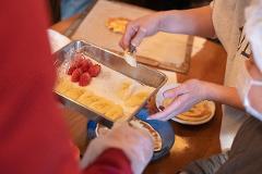 A person is carefully placing sliced bananas onto a plate of already made crepes, with strawberries also prepared for topping.