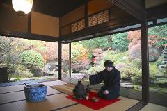 A man in a traditional robe kneels and performs a tea ceremony in a room overlooking a serene Japanese garden.