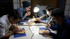 Visit Nikko's Craftsmanship Workshop to Experience Historic Techniques Four people carve wood on a table illuminated by a lamp, working on different pieces.