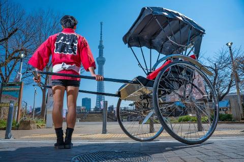 A rickshaw puller in traditional attire stands next to a rickshaw with the Tokyo Skytree in the background.