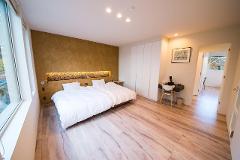A bright and airy bedroom features a large bed with white linens, a desk, and a view of a balcony through a doorway.