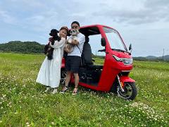 A couple holds two dogs in front of a red three-wheeled vehicle in a grassy field with a hill in the background.