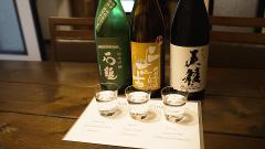 Three bottles of sake stand behind three shot glasses filled with sake on a wooden table, with a sake tasting menu in front.