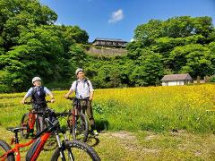 Two men in helmets and smart casual attire stand with their e-bikes in a field of yellow flowers, with a traditional Japanese building visible on a hill in the background.