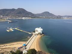 Swim with Dolphins in Kagawa! A serene view of a coastline with a unique white building on a peninsula, connected by a long, sandy path, set against a backdrop of rolling hills and calm, blue waters.