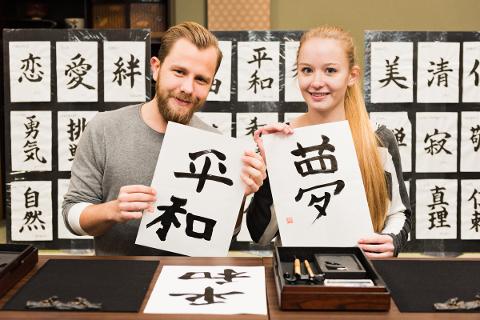 A smiling man and woman hold up Japanese calligraphy works featuring large black characters on white paper, with more calligraphy displayed behind them.