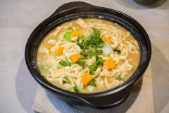 A steaming bowl of Japanese udon noodle soup with tofu, bok choy, and pumpkin is presented in a black ceramic pot on a light-colored table.