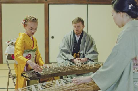 Two women in kimonos play kotos while a man in traditional Japanese clothing looks on in a room with tatami mats and shoji screens.
