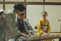 A woman in a green kimono teaches a man in a gray kimono how to play a koto, while another woman in a yellow kimono watches with a smile.