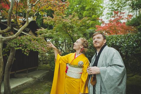 A couple in traditional Japanese attire, a woman in a yellow kimono and a man in a grey robe, admire the autumn foliage in a Japanese garden.