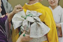 A woman wearing a yellow kimono has her obi sash adjusted by another woman in a purple kimono, while a third woman in a white kimono stands nearby.