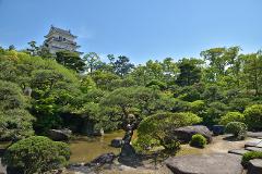 A serene Japanese garden features lush greenery, a small pond, and a traditional castle nestled among the trees under a clear blue sky.