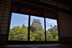 A majestic Japanese castle is framed by a traditional wooden window, surrounded by lush green trees under a blue sky.