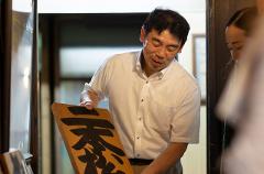 A man shows a wooden sign with Japanese characters to someone out of frame in front of a doorway in a traditional building.