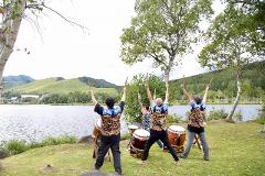 A group of taiko drummers perform by a lake with rolling hills in the background.