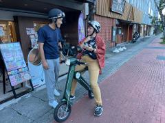 A man and a woman wearing helmets stand next to a green electric scooter on a sidewalk in front of shops.