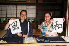 A man and a woman, both dressed in traditional Japanese clothing, hold up pieces of paper with Japanese calligraphy on them.