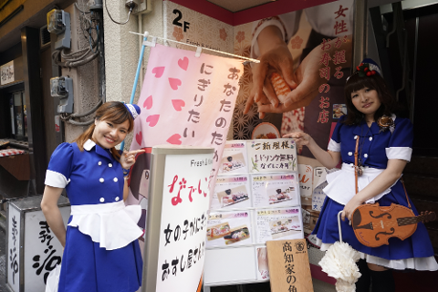 Two women in blue maid costumes pose outside a Japanese restaurant with menu boards and a pink banner advertising sushi.