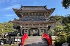 A majestic, multi-tiered Japanese temple gate stands grandly under a clear blue sky, with a vibrant red arched bridge leading towards its entrance.