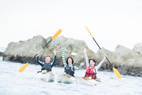 Three women in kayaks raise their arms and paddles in front of a rocky shoreline, smiling and celebrating on the water.