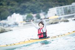A smiling woman in a red life vest sits in a yellow kayak, raising her paddle in the air in front of a blurred background of water, rocks, and buildings.