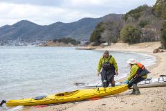 Two people in kayaking gear pull a yellow kayak onto a sandy beach, with other kayaks and mountains in the background.