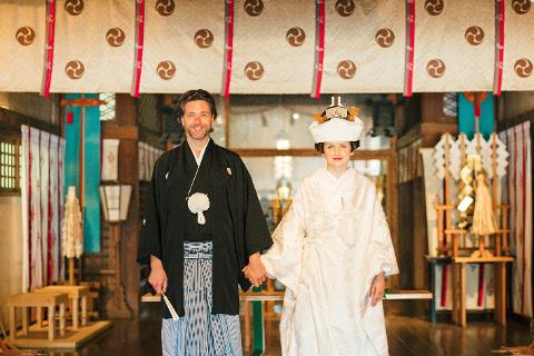 Kanazawa 5H Wedding Experience with Parade & Kaiseki Meal A smiling groom in a black kimono and a bride in a white wedding kimono stand hand-in-hand in front of a decorated backdrop.