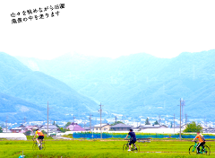 Three cyclists ride through a green field with mountains in the background.