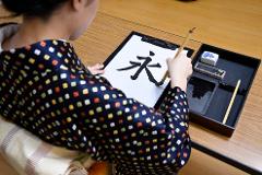 A person in a patterned kimono practices Japanese calligraphy, writing the character "永" on white paper.