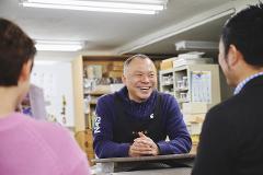A smiling man wearing an apron and hoodie sits at a table talking with two people in a workshop setting.