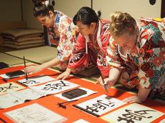 Three women in kimonos are practicing Japanese calligraphy on large sheets of paper spread on a tatami mat floor.